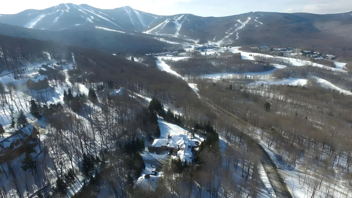 A secluded house nestled in a dense forest, surrounded by snow-covered mountains and trees. The wintry landscape highlights ski trails and invites thoughts of outdoor adventures and mountain tranquility. The house is set against a stunning mountain backdrop, encapsulating the essence of slopeside living.