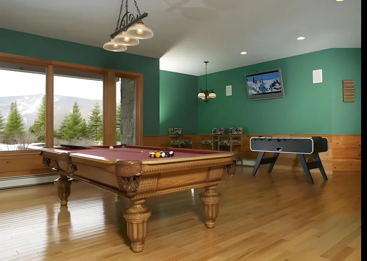 A lively game room featuring a pool table, foosball table, large windows with mountain views, chandeliers, and a cozy seating area. The space is designed for both relaxation and entertainment, with natural light and inviting decor.