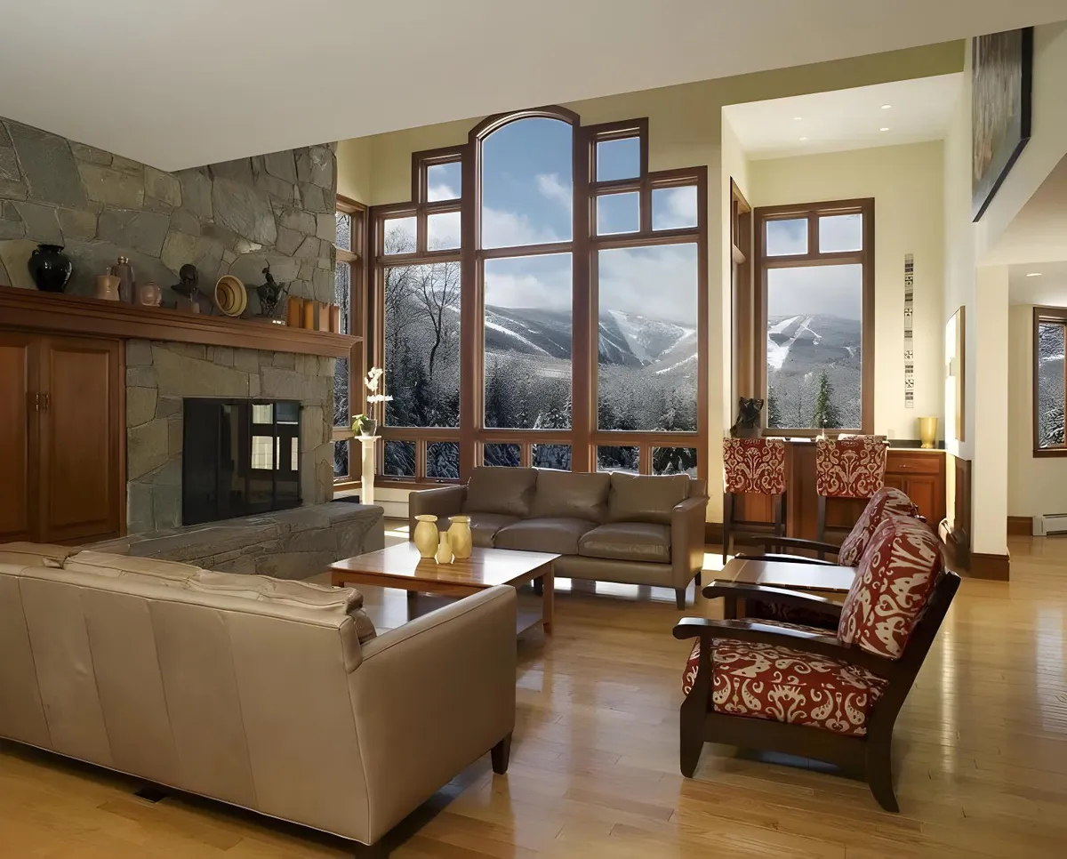 A spacious great room featuring a prominent stone wood-burning fireplace, comfortable seating arranged to take advantage of both the warmth and the panoramic mountain views through expansive windows. The natural wood finishes and cozy furnishings set a relaxing, inviting scene ideal for après-ski gatherings or family relaxation.