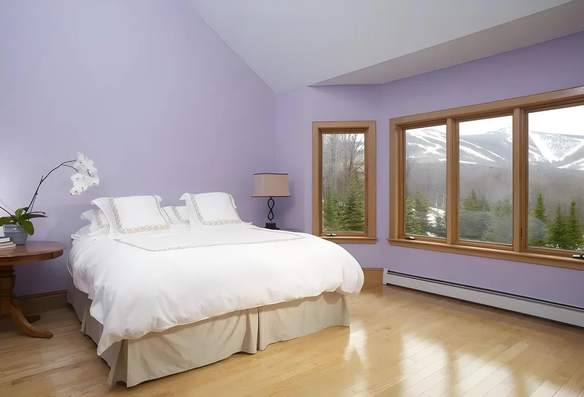 A spacious bedroom featuring a large bed with crisp white bedding, flanked by a bedside lamp and a delicate orchid on the table. The room is illuminated by natural light pouring through large windows that frame a serene mountain view, creating an inviting and tranquil atmosphere.