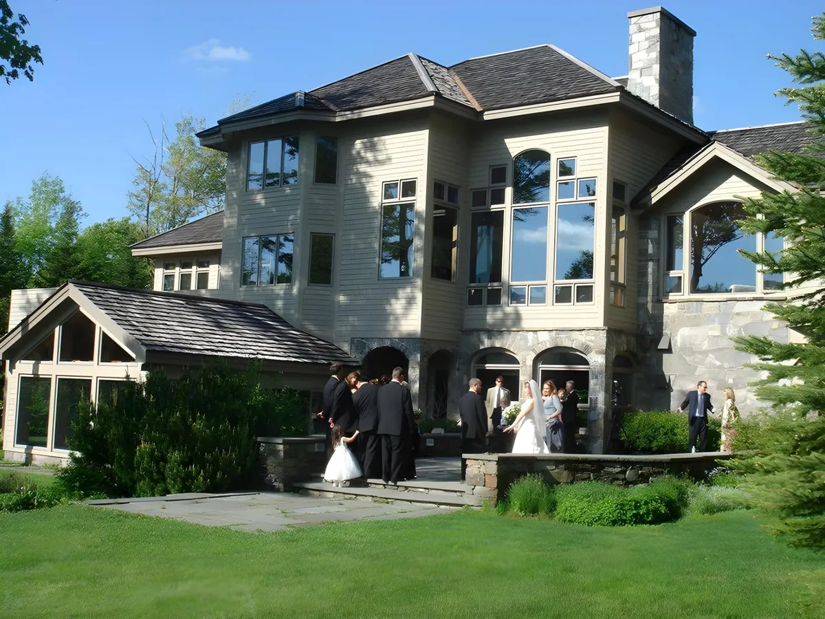A grand, luxurious mountainside house with expansive windows serves as the backdrop to an elegant outdoor wedding ceremony. Joyful guests in formal attire celebrate amidst stunning wedding decorations, while the picturesque Vermont landscape surrounds the scene.